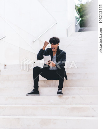 Modern cheering African American man using tablet while sitting at urban staircase on street 108213365