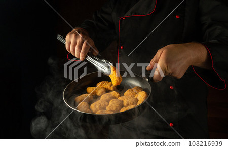 The cook prepares chicken nuggets in a frying pan. The concept of cooking nagits in the kitchen of a restaurant or hotel. Free space for advertising on a dark background The cook prepares chicken nuggets in a frying pan. The concept of cooking nagits in the kitchen of a restaurant or hotel. Free space for advertising on a dark background 108216939