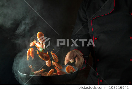 The cook cooks shrimp in a hot pan with steam. The concept of cooking seafood or healthy vegetarian food and eating on a dark background. Freeze in motion. Free space for recipe. 108216941
