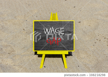 Wage gap symbol. Concept words Wage gap on wooden blocks on a beautiful black chalk blackboard on a beautiful beach sand background. Business, support and wage gap concept. Copy space. Wage gap symbol. Concept words Wage gap on wooden blocks on a beautiful black chalk blackboard on a beautiful beach sand background. Business, support and wage gap concept. Copy space. 108218298