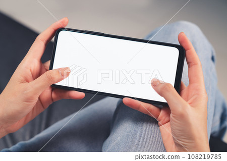 Hand holding black smartphone with blank screen for graphic display montage in the living room Hand holding black smartphone with blank screen for graphic display montage in the living room 108219785