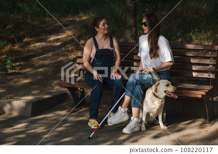 Blind caucasian woman sitting on bench with guide dog and pregnant friend.  108220117