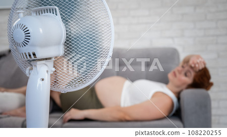 Pregnant woman suffers from heat and lies on the sofa near the electric fan.  108220255