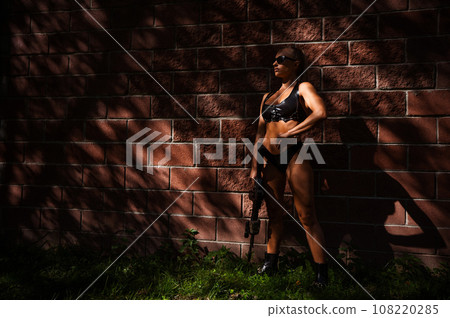 A caucasian woman in a bikini holds a machine gun against a brick wall. A caucasian woman in a bikini holds a machine gun against a brick wall. 108220285