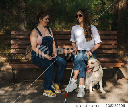 Blind caucasian woman sitting on bench with guide dog and pregnant friend. Blind caucasian woman sitting on bench with guide dog and pregnant friend. 108220306