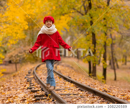 Caucasian girl in a red coat and beret walks along the railway tracks in the park in autumn. 108220389