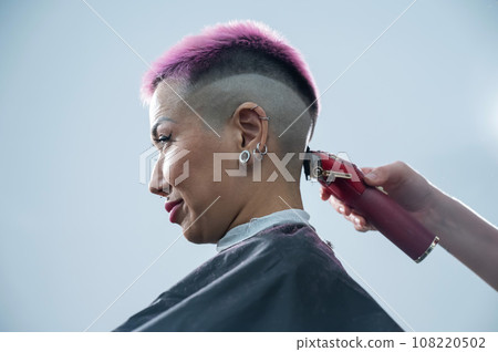 The hairdresser shaves the back of the head of a female client. Rear view of a woman with short pink hair in a barbershop. The hairdresser shaves the back of the head of a female client. Rear view of a woman with short pink hair in a barbershop. 108220502