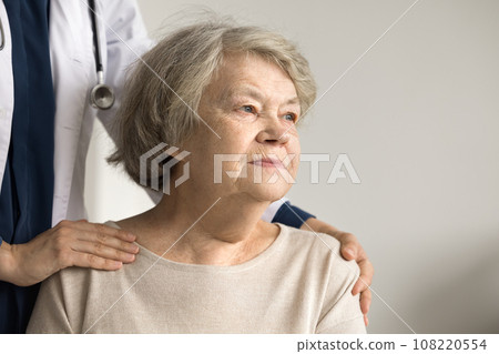 Older woman looking into distance, therapist touch shoulders supporting patient Older woman looking into distance, therapist touch shoulders supporting patient 108220554
