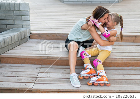 Little girl in roller skates and her mom sit on a wooden ladder and hug outdoors. Little girl in roller skates and her mom sit on a wooden ladder and hug outdoors. 108220635