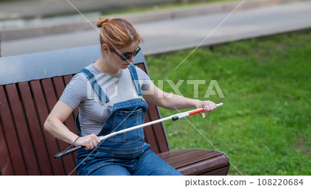 Blind pregnant woman takes out a folding cane while sitting on a bench.  108220684