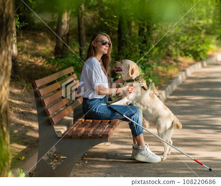 Blind young woman cuddling with guide dog while sitting on a bench. Blind young woman cuddling with guide dog while sitting on a bench. 108220686