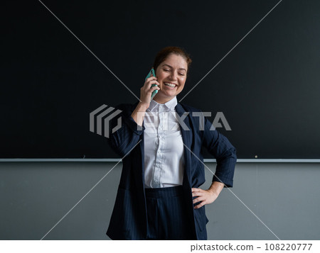 Caucasian female teacher in a suit talking on the phone while standing at the blackboard. Caucasian female teacher in a suit talking on the phone while standing at the blackboard. 108220777