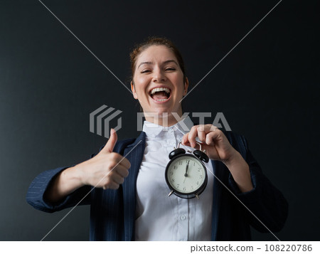 Happy female teacher in pantsuit holding an alarm clock standing at the school board and showing thumbs up. Happy female teacher in pantsuit holding an alarm clock standing at the school board and showing thumbs up. 108220786
