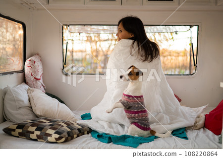 Caucasian woman looks out of the window of the van while hugging her dog Jack Russell Terrier. Travel in a camper in autumn. Caucasian woman looks out of the window of the van while hugging her dog Jack Russell Terrier. Travel in a camper in autumn. 108220864