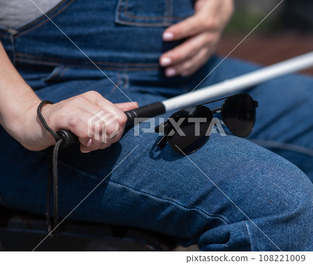 A blind pregnant woman sits on a bench and holds a folding tactile cane in her hands. A blind pregnant woman sits on a bench and holds a folding tactile cane in her hands. 108221009