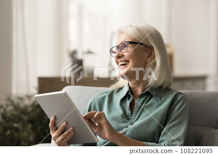 Cheerful elderly woman resting on home sofa with tablet Cheerful elderly woman resting on home sofa with tablet 108221095