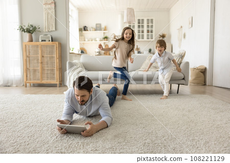 Dad rest on floor use tablet while children play nearby Dad rest on floor use tablet while children play nearby 108221129