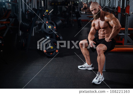 A muscular bald man in shorts is resting on a bench after a workout. Bodybuilder showing off his shape in the gym. A muscular bald man in shorts is resting on a bench after a workout. Bodybuilder showing off his shape in the gym. 108221163