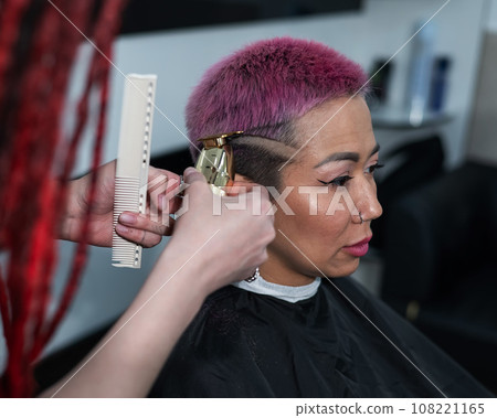 The hairdresser shaves the temple of a female client. Asian woman with short pink hair in barbershop. The hairdresser shaves the temple of a female client. Asian woman with short pink hair in barbershop. 108221165