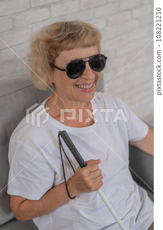 An elderly blind woman wearing sunglasses and with a tactile cane sits on the sofa.  108221210