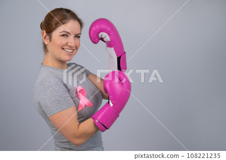 Caucasian woman in pink boxing gloves with a pink ribbon on her chest on a gray background. Fight against breast cancer.  108221235