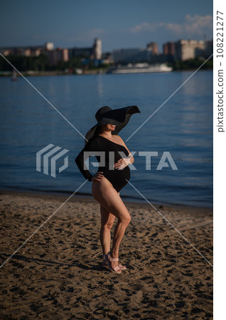Pregnant woman in a large straw hat and black swimsuit posing on the beach. 108221277