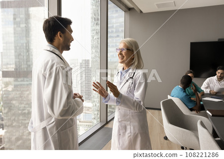 Happy confident mature doctor woman speaking to younger practitioner Happy confident mature doctor woman speaking to younger practitioner 108221286