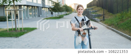 A woman rides an electric scooter with a dog in a backpack. Pappilion Spaniel Continental in a sling. Widescreen.  108221353