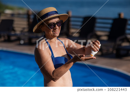 Portrait of an old woman in a straw hat, sunglasses and a swimsuit applying sunscreen to her skin while relaxing by the pool.  108221443