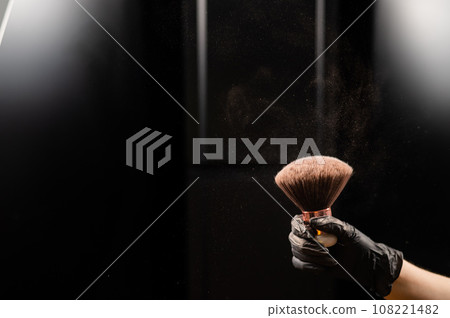 A woman shakes a large powder brush with clouds of dust on a black background. Treatment after instant tanning A woman shakes a large powder brush with clouds of dust on a black background. Treatment after instant tanning 108221482