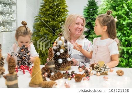Elderly caucasian woman making pine cones decoration for christmas with two granddaughters 108221568