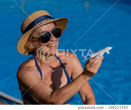 An elderly woman in a hat and sunglasses holds a model airplane while sitting by the pool. Retirement vacation concept. An elderly woman in a hat and sunglasses holds a model airplane while sitting by the pool. Retirement vacation concept. 108221593