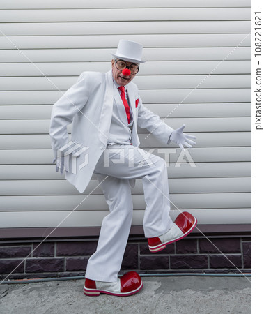 An elderly man in a white suit, huge boots and a clown nose walks funny. An elderly man in a white suit, huge boots and a clown nose walks funny. 108221821