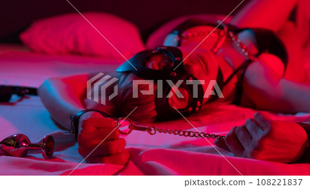 Sexy blonde woman in a leather mask and handcuffs and a collar lies in the bedroom in a red-blue neon light. Role-playing games. 108221837