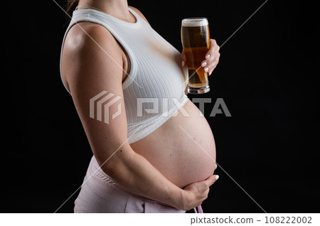 Close-up of the belly of a pregnant woman holding a glass of beer on a black background. Skin rash. Close-up of the belly of a pregnant woman holding a glass of beer on a black background. Skin rash. 108222002