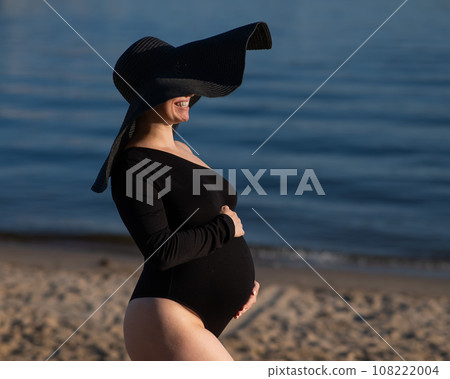 Pregnant woman in a large straw hat and black swimsuit posing on the beach. Pregnant woman in a large straw hat and black swimsuit posing on the beach. 108222004