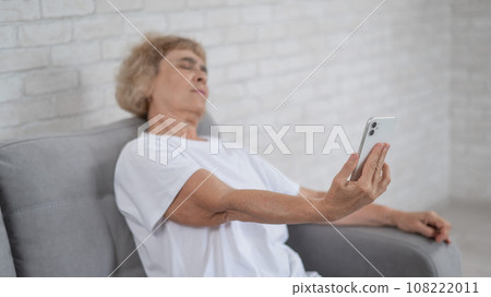 An elderly Caucasian woman suffers from farsightedness and tries to read a smartphone with her arm outstretched.  108222011