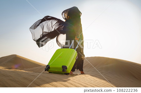 Arabian woman in traditional abaya costume with bright light green or yellow suitcase in sands. Concept and idea of travel to United Arab Emirates, sand dunes in desert of Dubai at sunset. 108222236