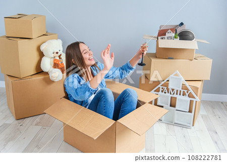 Happy woman drinks champagne from a glass while sitting inside a cardboard box in new apartment. Happy woman drinks champagne from a glass while sitting inside a cardboard box in new apartment. 108222781