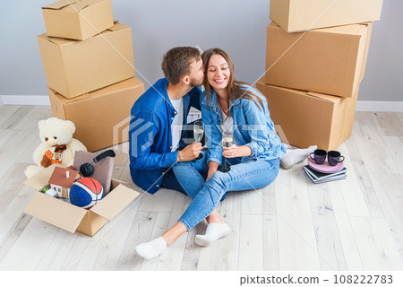 Couple taking a break on moving day, sitting on floor in new apartment celebrating with champagne surrounded by cardboard boxes. Couple taking a break on moving day, sitting on floor in new apartment celebrating with champagne surrounded by cardboard boxes. 108222783