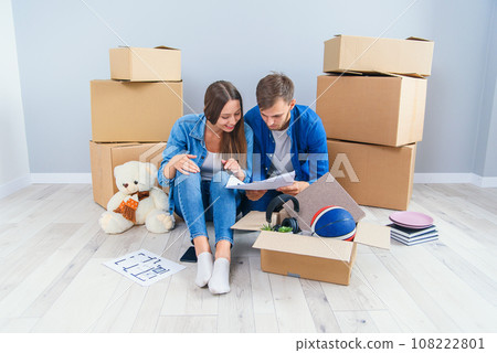 Couple have a discussion about design of their new home sitting on the wooden floor between many cardboard boxes. Beautiful young stylish couple in love planning home design for their new bought flat. Couple have a discussion about design of their new home sitting on the wooden floor between many cardboard boxes. Beautiful young stylish couple in love planning home design for their new bought flat. 108222801