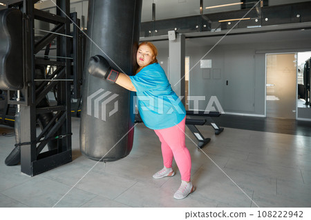 Tired exhausted fat woman leaning and hugging punching bag while training in gym Tired exhausted fat woman leaning and hugging punching bag while training in gym 108222942