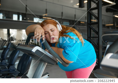 Cute young woman with obesity falling asleep on treadmill during training Cute young woman with obesity falling asleep on treadmill during training 108222945