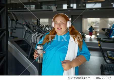 Portrait of young overweight woman wearing sportive clothes and headphones at gym Portrait of young overweight woman wearing sportive clothes and headphones at gym 108222946