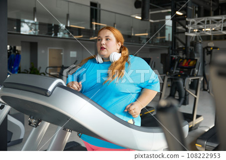 Active overweight woman running on treadmill track enjoying cardio exercise Active overweight woman running on treadmill track enjoying cardio exercise 108222953
