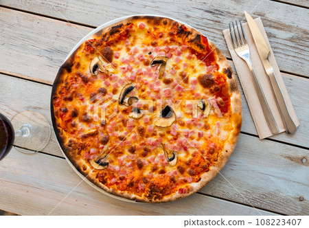 On plate is small round pizza with cubes of ham and slices of mushrooms 108223407