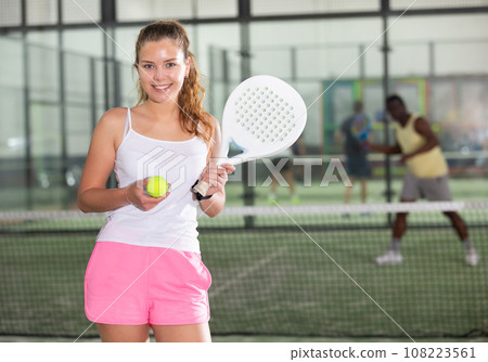 Smiling girl with paddle tennis racquet and ball standing on indoor court Smiling girl with paddle tennis racquet and ball standing on indoor court 108223561