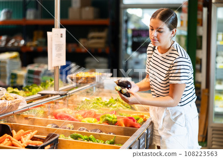 Positive girl choosing fresh eggplants in vegetables department of supermarket Positive girl choosing fresh eggplants in vegetables department of supermarket 108223563