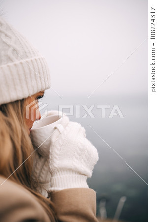 Young pretty woman holding cup of tea enjoying winter frozen nature by the sea 108224317