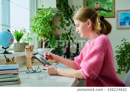 Teenage girl student illustrator drawing on digital tablet sitting at desk at home 108224682